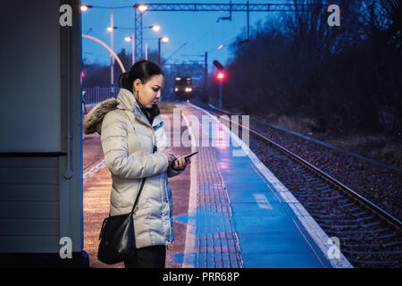 A woman is wating for a train and uses a smartphone at a railway station on the background of a railway and a catchy train at night, Glasgow, Scotland Stock Photo