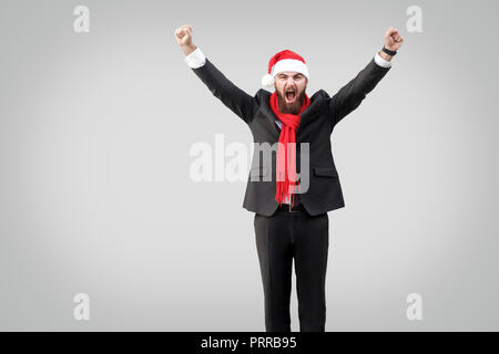 Bearded businessman in classic black suit, red scarf and new year hat, looking at camera with raised arms and screaming. Wining and success concept. I Stock Photo