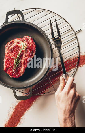 cropped image of man holding meat fork and cooking steak with rosemary in kitchen Stock Photo