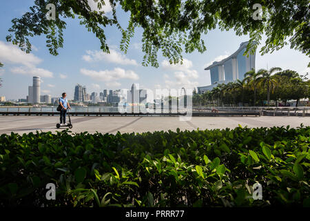 Through the foliage, a man on his electric mobility scooter riding past the backdrop of skyscrapers and Marina Bay Sands hotel. Singapore. Stock Photo