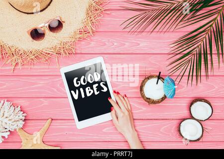 cropped shot of woman using tablet with 'good vibes' inspection on pink wooden surface with coconut cocktail and straw hat Stock Photo