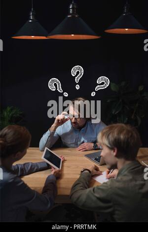 Businesswoman showing something on tablet to her partners with question marks over head and sitting at table Stock Photo