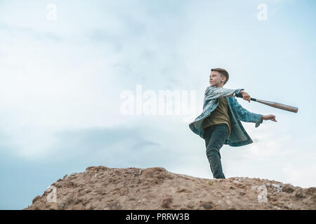 low angle view of boy in denim jacket playing with baseball bat while standing on hill against cloudy sky Stock Photo