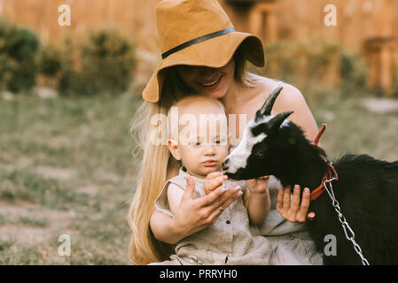 portrait of smiling mother in hat and little son petting domestic goat at countryside Stock Photo