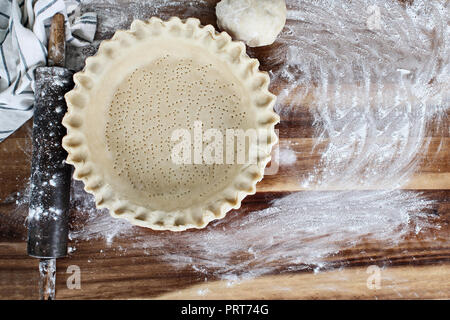 Homemade butter pie crust in pie plate with fluted pinched edge, rolling pin, towel and extra ball of dough over floured rustic wooden background. Stock Photo