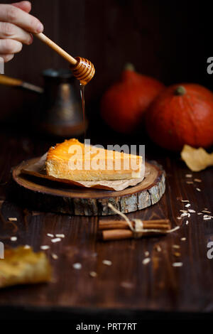 Female hand pours honey pumpkin cheesecake. Pumpkins, coffee, foliage, vanilla on a wooden dark background. Autumn and winter cozy concept Stock Photo