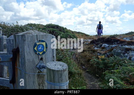 Lone Woman Hiker Walking past Wooden Gate & Signpost for the Isle of Anglesey Coastal Path near the Fishing Village of Cemaes, Wales, UK. Stock Photo