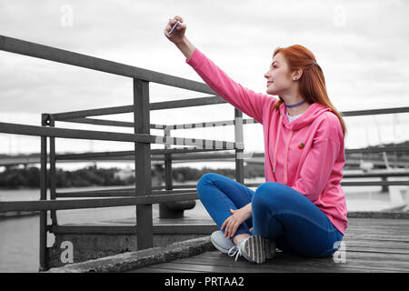 Red haired girl smiling while taking selfies on the pier Stock Photo