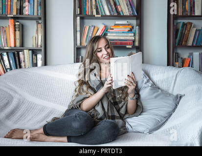 the girl with long hair dressed comfortably reading a book on the couch in the background of the library, the concept of reading and leisure time week Stock Photo