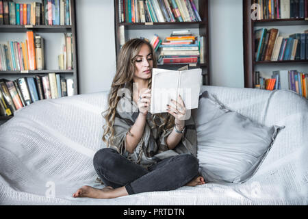 the girl with long hair dressed comfortably reading a book on the couch in the background of the library, the concept of reading and leisure time week Stock Photo