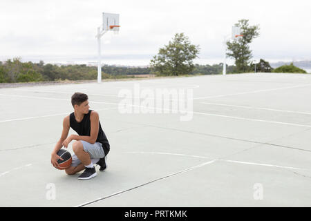 Male teenage basketball player crouching with ball on basketball court Stock Photo