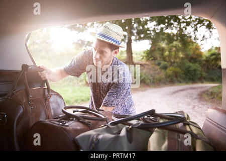 Young man in trilby removing luggage from car boot on rural road Stock Photo