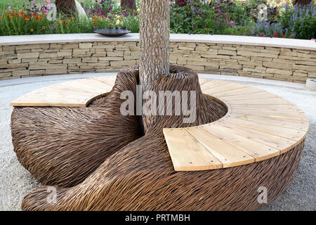 Garden with a Circular patio design with Woven willow and wooden bench seating under tree Cotswold dry stone wall bench patio seating area UK summer Stock Photo