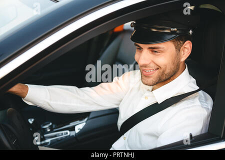 Portrait of young joyful man taxi driver in uniform and cap driving car wearing seat belt Stock Photo