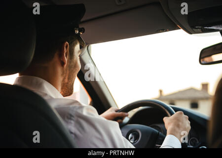 Portrait from back of caucasian chauffeur man wearing uniform and cap holding wheel and driving car Stock Photo