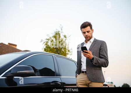 Portrait of rich businessman wearing suit, standing near his luxury black car and using smartphone while holding in hand
