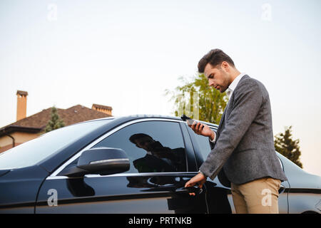 Portrait of caucasian businessman wearing suit, opening driver's door of his luxury black car and using smartphone Stock Photo