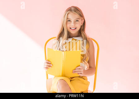 smiling fashionable kid reading book while sitting on yellow chair on pink Stock Photo