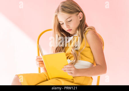 adorable elegant kid reading book while sitting on yellow chair on pink Stock Photo
