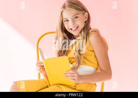 happy stylish child reading book while sitting on yellow chair on pink Stock Photo