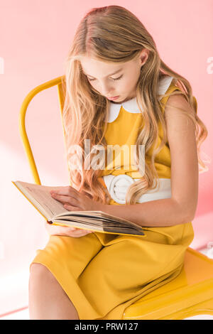 stylish blonde youngster reading book while sitting on yellow chair on pink Stock Photo