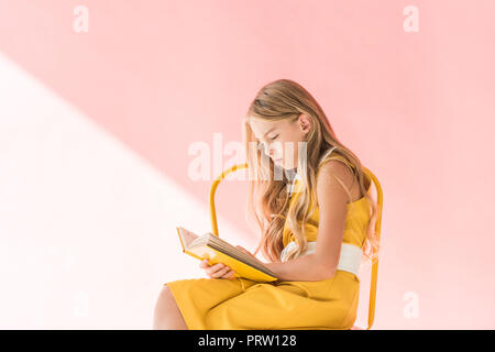 stylish adorable child reading book while sitting on yellow chair on pink Stock Photo