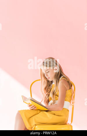 stylish blonde child reading book while sitting on yellow chair on pink Stock Photo