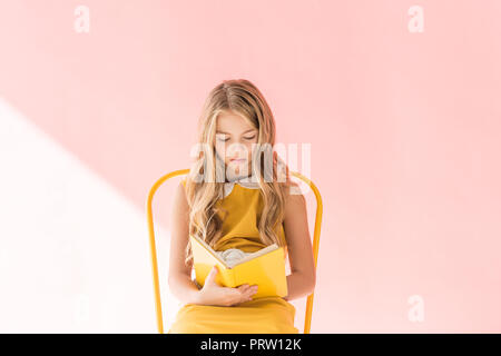 beautiful blonde youngster reading book while sitting on yellow chair on pink Stock Photo