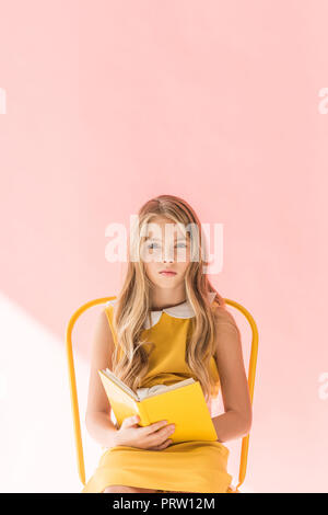 fashionable youngster reading book while sitting on yellow chair on pink Stock Photo