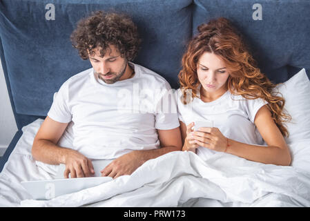 high angle view of young couple using laptop and smartphone in bed Stock Photo