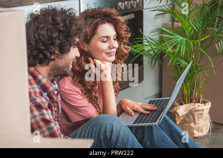 side view of cheerful couple sitting with laptop near cardboard boxes on floor in new kitchen Stock Photo