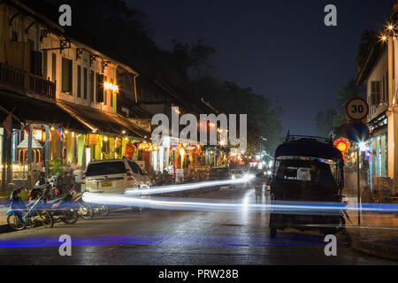 Scooters, cars and three-wheeler taxis (jumbo or tuk-tuk), several people, lit colonial era buildings and cars' light trails in Luang Prabang at dusk. Stock Photo