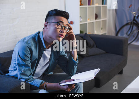 thoughtful young asian man in eyeglasses holding notebook and looking away at home Stock Photo