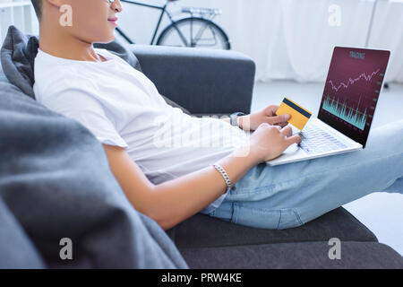 cropped image of man using laptop with traiding appliance and holding credit card on sofa at home Stock Photo