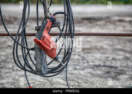 Close-up of a new modern red drill with a long wire wrench attachment hanging ready for work against a building site Stock Photo