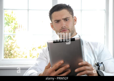 Handsome male doctor, gp, wearing white lab coat, holding and studying digital tablet Stock Photo