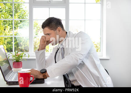 Handsome, male doctor wearing white lab coat at desk looking at laptop computer with window in background Stock Photo