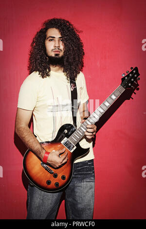 Closeup of one handsome passionate expressive cool young brunette  rock musician men with long curly hair playing electrIC guitar standing against red Stock Photo