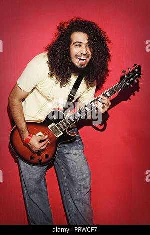 Closeup of one handsome passionate expressive cool young brunette rock musician men with long curly hair playing electric guitar standing against red Stock Photo