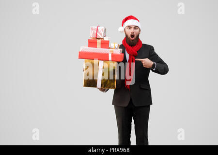 Surprised handsome businessman with beard, black suit, red scarf and new year hat holding, pointing gift box and looking at camera with shocked face.  Stock Photo