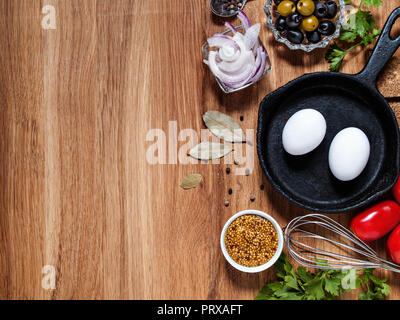 On a wooden background horizontally with copyscera chicken eggs in a cast iron pan, onions, olives, tomatoes, greens, a beater Stock Photo
