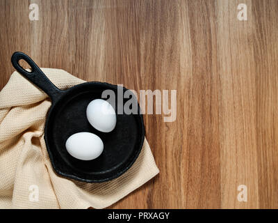 A black cast-iron pan with chicken eggs stands on a beige fabric napkin on a wooden background horizontally with copy space Stock Photo