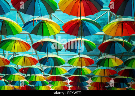 Colorful display of open rainbow colored umbrellas outdoors in street under blue awning on sunny day. Stock Photo