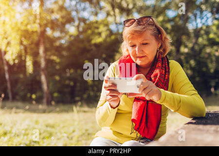 Middle-aged woman relaxing using phone outdoors. Stylish senior lady watching video on smartphone in autumn forest. Stock Photo