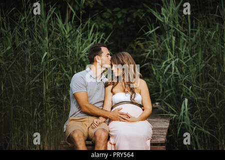 Pregnant woman and her husband are sitting at a lake while he is kissing her forehead gently Stock Photo