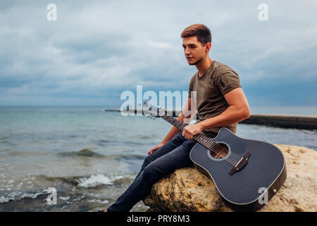 Upset young man holding acoustic guitar on beach surrounded with rocks on rainy day. Feeling lonely and sad. Depression Stock Photo