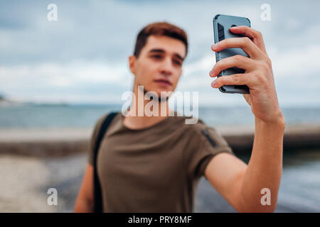 Young man taking selfie using phone on cloudy beach. Handsome sportive guy walking by pier in Odesa Stock Photo