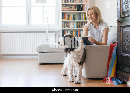 Smiling mature woman with dog lying on couch at home holding tablet Stock Photo