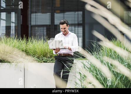 Smiling businessman using laptop outside office building Stock Photo
