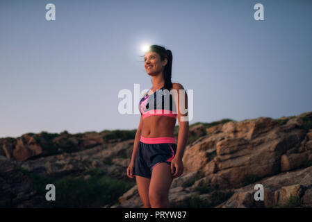 Sportive woman with headlamp standing on rocky coast Stock Photo